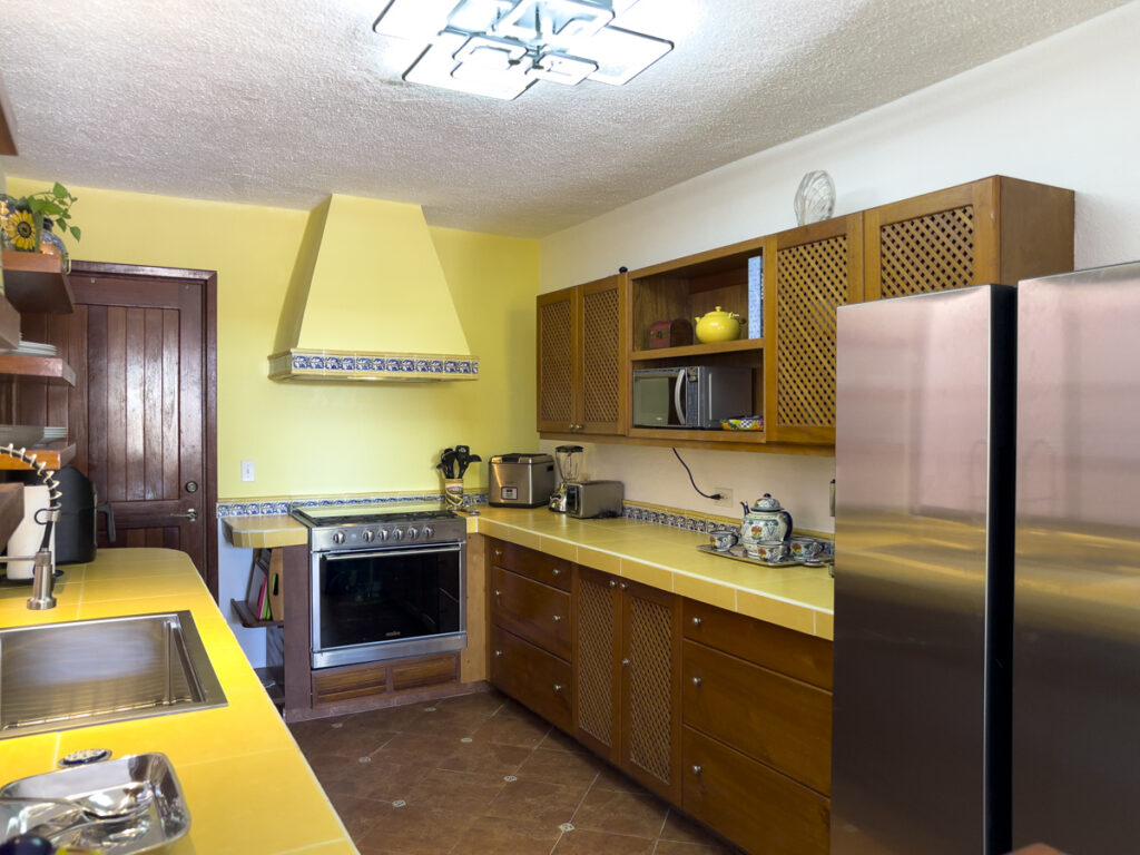 Wooden kitchen cabinets and yellow countertops beside a stove, hood, and stainless steel refrigerator.