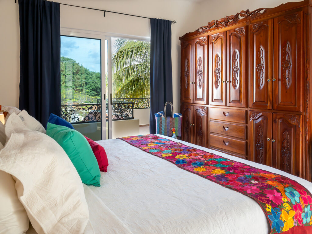 Bed with a floral runner facing a carved wooden wardrobe beside balcony doors overlooking palms.