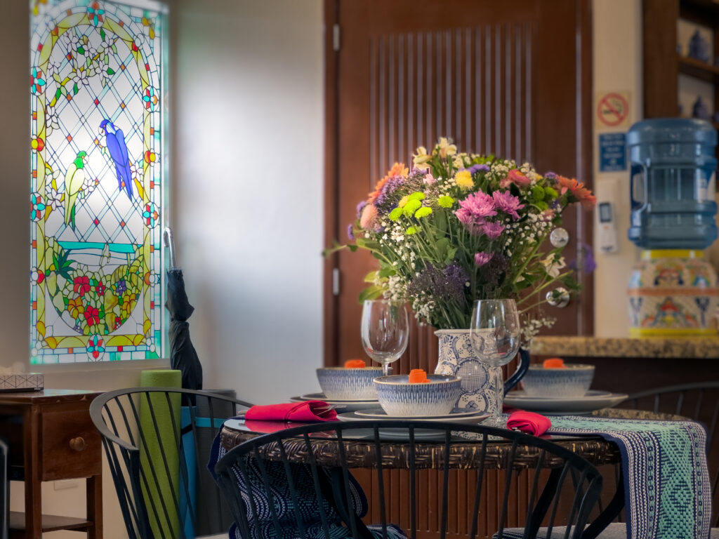 Flower arrangement and place settings on a dining table beside a colorful stained-glass panel.