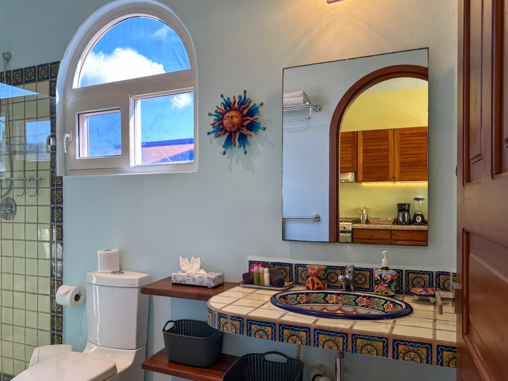 Decorated tiled sink and counter beneath an arched mirror beside a toilet and window.