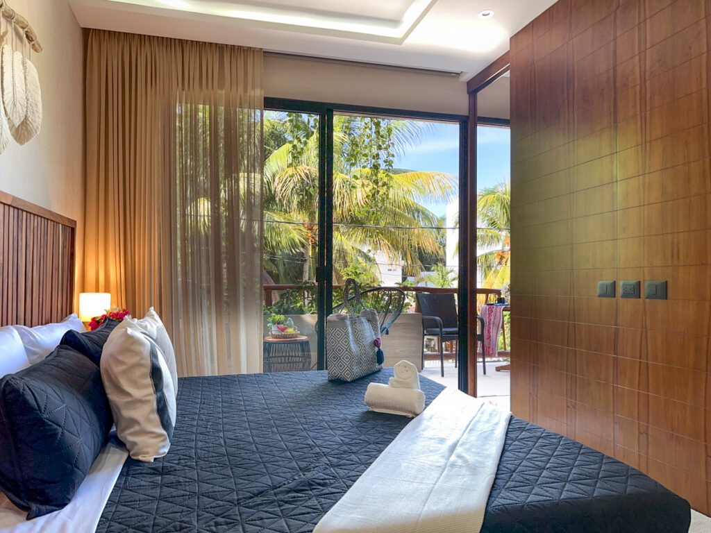 Bed with dark bedding facing large glass doors opening to a tree-lined balcony.