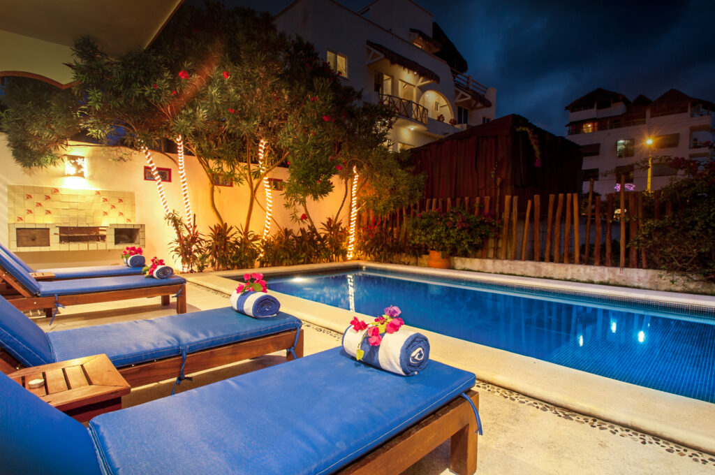 Blue-lit swimming pool beside lounge chairs with rolled towels and flowers at night.