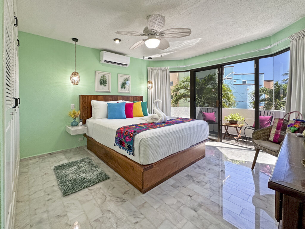 Bed with bright pillows and a floral runner beside broad glass doors opening onto a balcony.