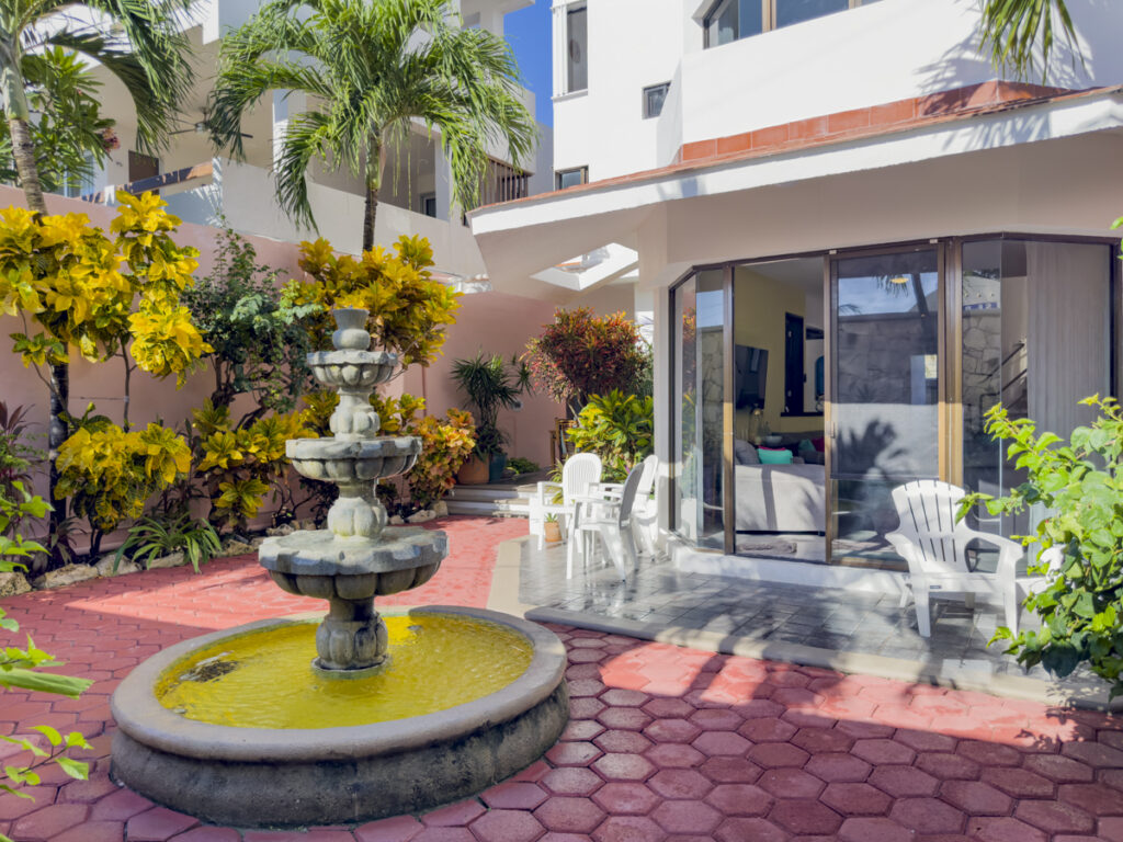 Courtyard fountain in front of open glass doors and patio chairs surrounded by tropical plants.