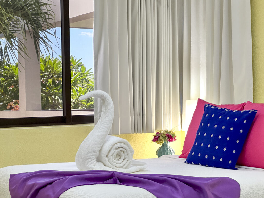 White towel swan on a bed with a purple throw beside bright pillows and a curtained window.