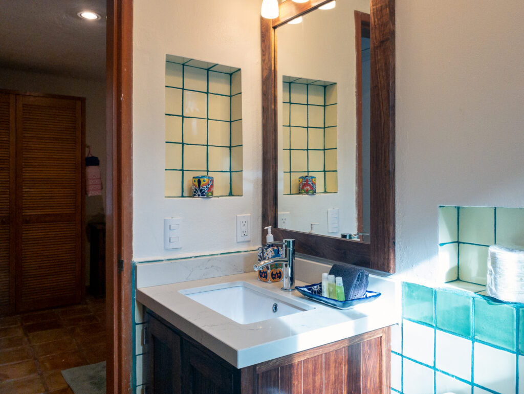 Rectangular basin and wooden vanity beneath a framed mirror beside green-bordered tiles.