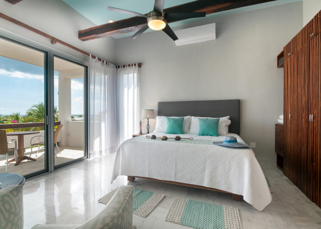 Bed with turquoise accents beneath dark ceiling beams beside a balcony table and chairs.