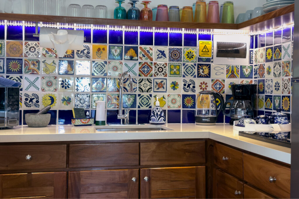 Kitchen sink and wooden cabinets beneath a multicolored tile backsplash and shelves of mugs.