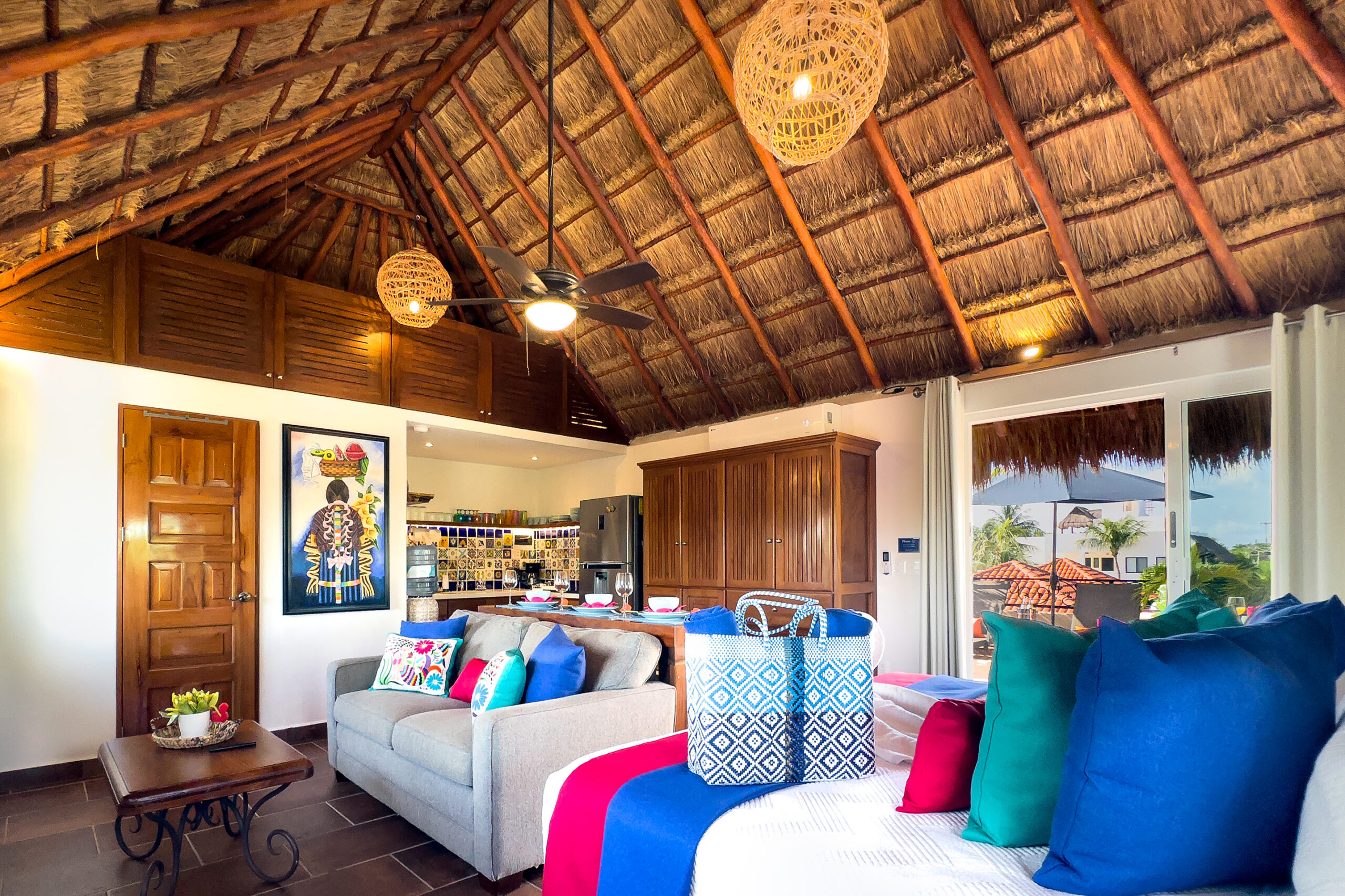 Colorful bed and gray sofa beside the kitchen beneath a high palapa roof.