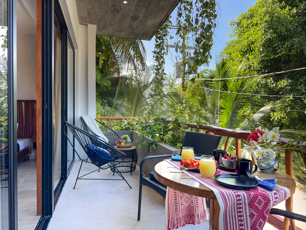 Table set with drinks and dishes beside balcony chairs and a railing surrounded by greenery.