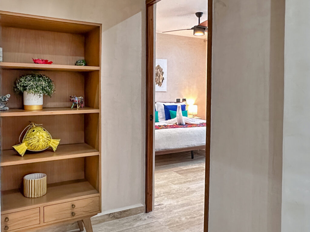 Open wooden display shelves beside a doorway looking into a bedroom with colorful bedding.