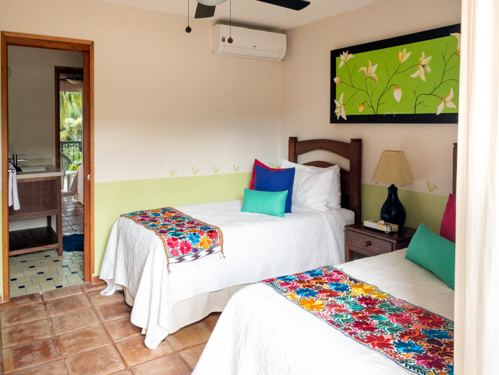 Two beds with colorful pillows and floral runners beneath green floral artwork.