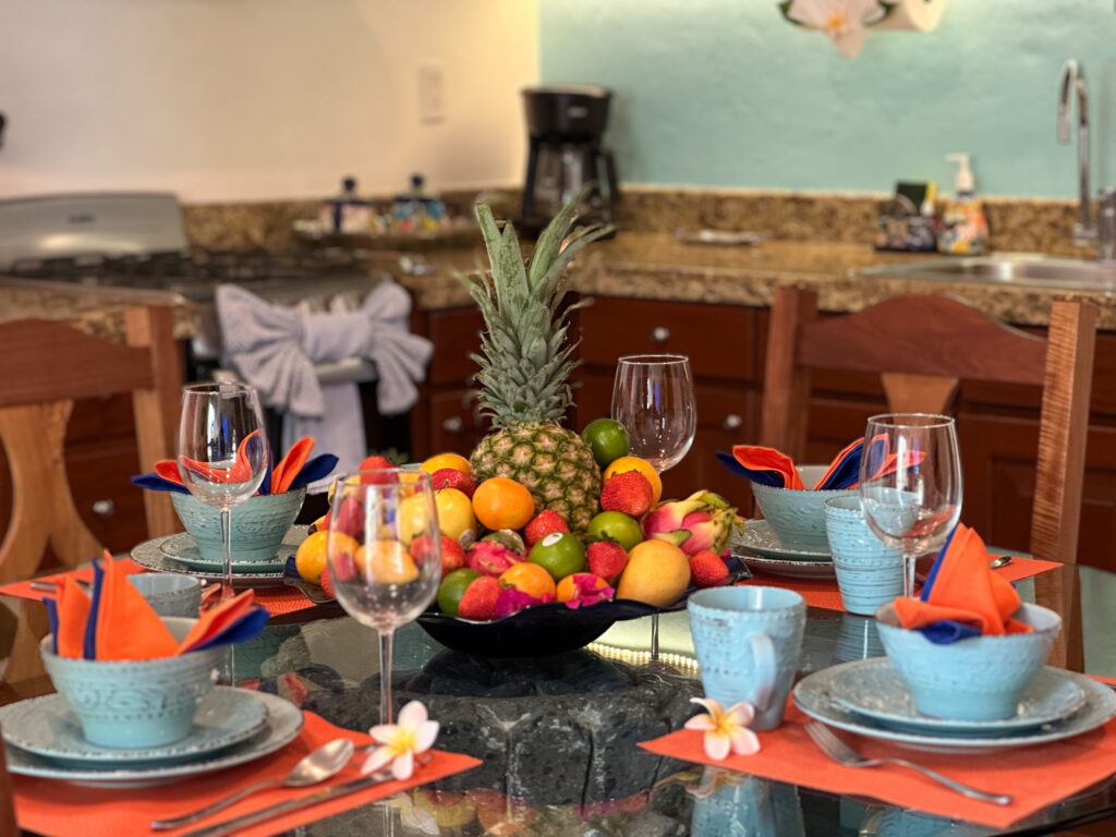 Bowl of tropical fruit and pineapple on a table set with blue dishes and orange napkins.