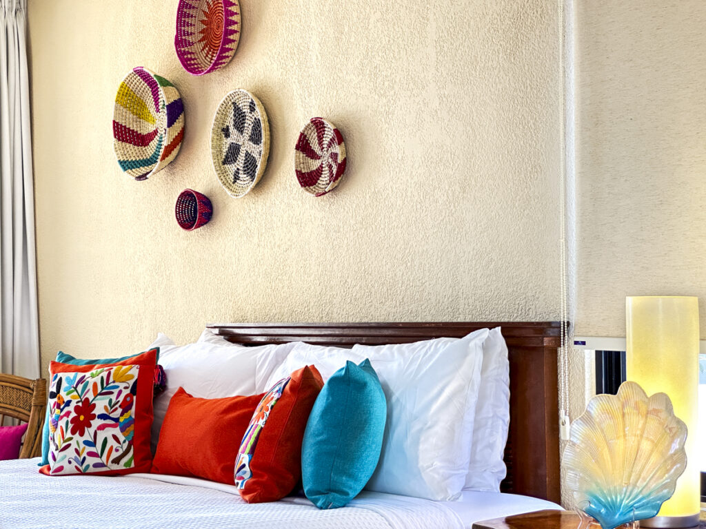 Colorful pillows on a bed beneath woven wall decorations and beside a shell-shaped lamp.
