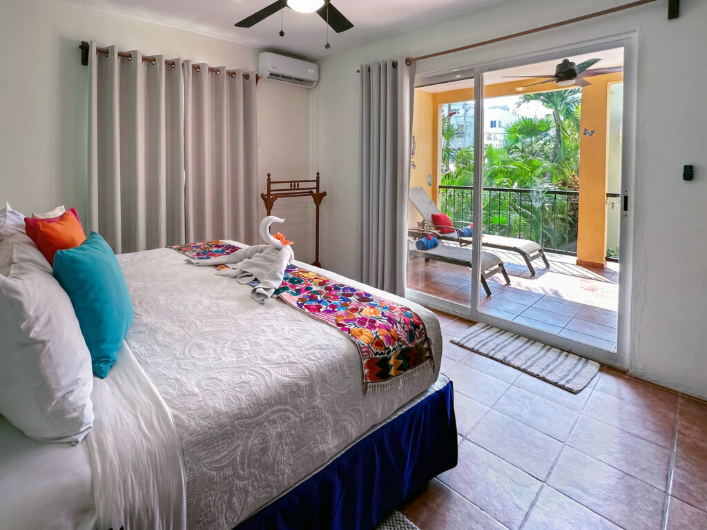Bed with a floral runner and towel swan beside sliding doors opening to balcony lounge chairs.