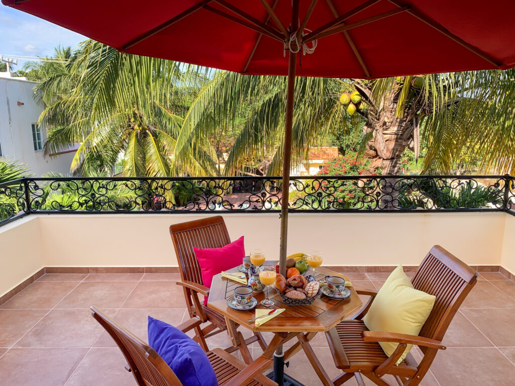 Wooden dining table and chairs with bright cushions beneath a red umbrella overlooking palms.