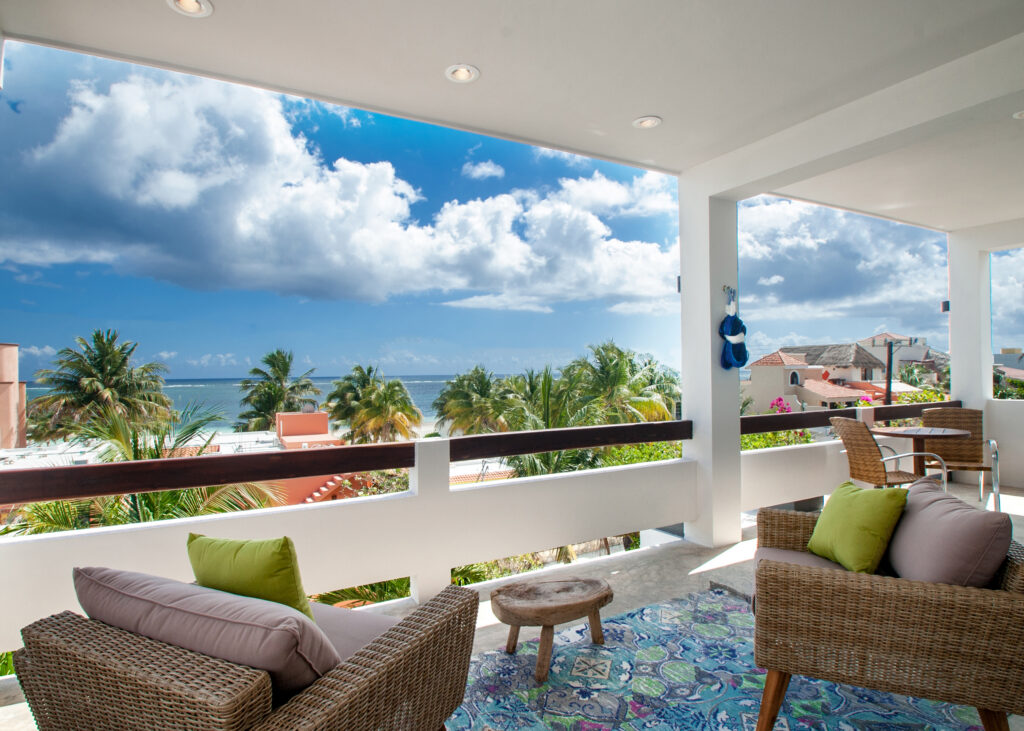 Woven lounge chairs and small wooden tables on a covered terrace overlooking palms and the coast.