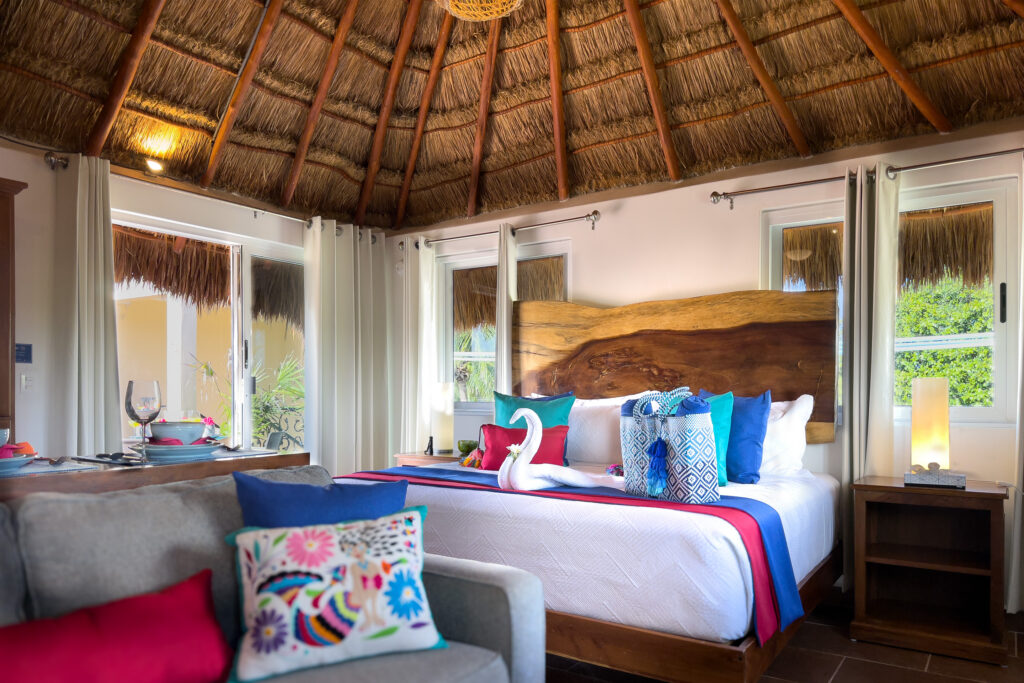 Bed with a wide natural wood headboard beside a gray sofa beneath exposed thatch and timber rafters.