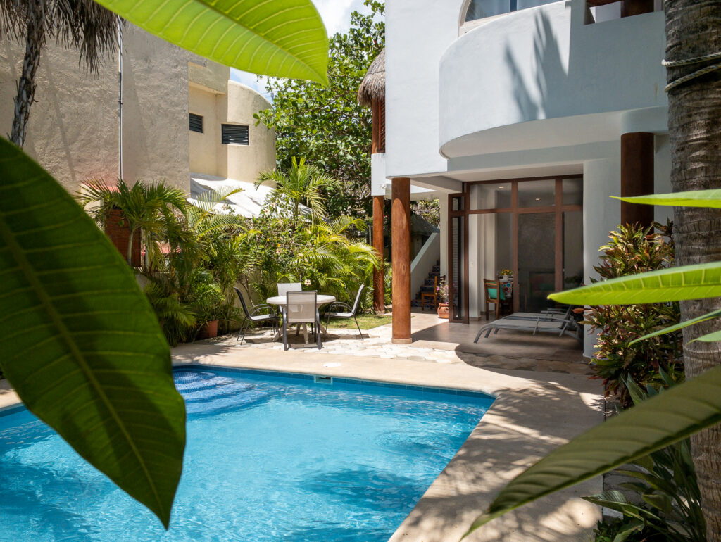 Pool framed by tropical foliage and a white building with covered patios.