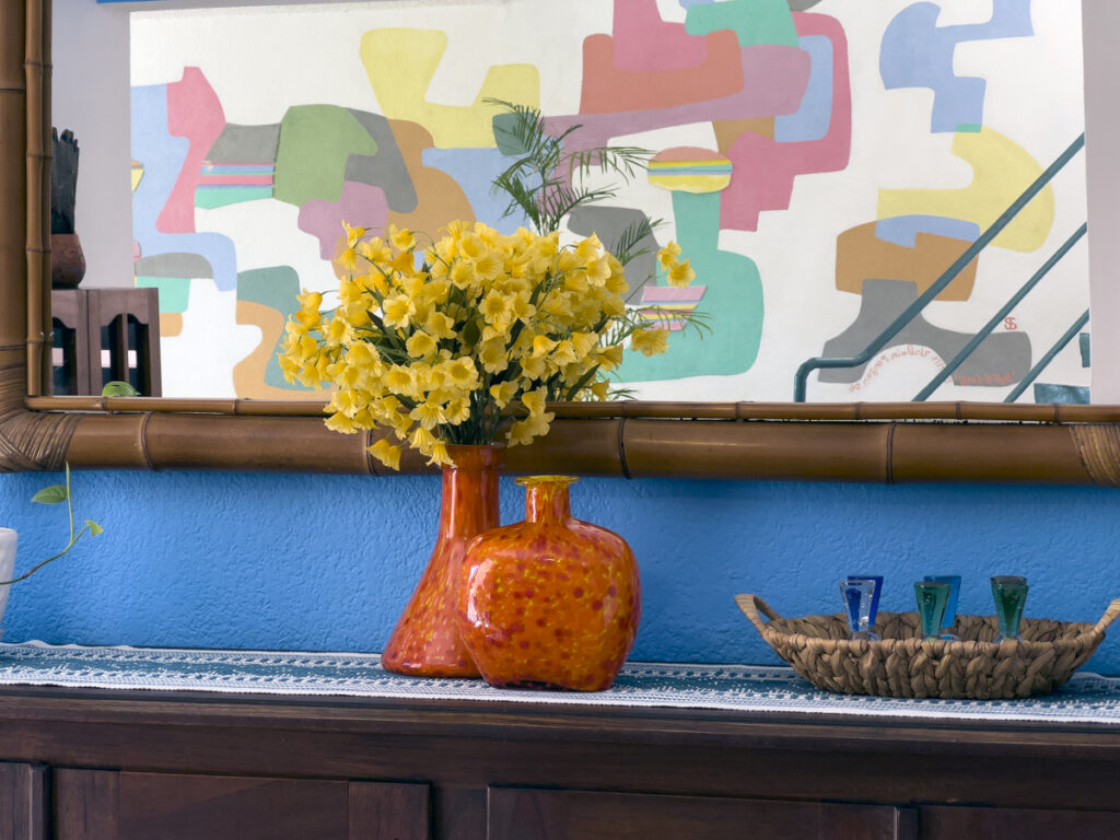 Yellow flowers in an orange vase beneath a mirror reflecting colorful abstract wall art.