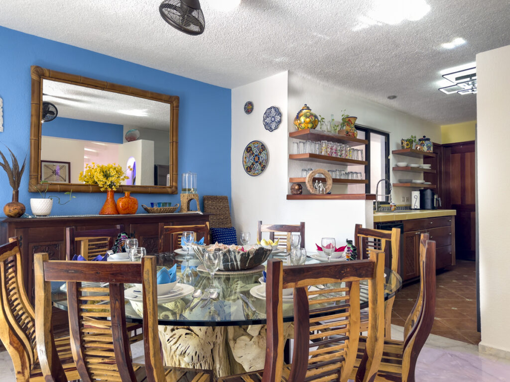 Dining table with wooden chairs near a sideboard, mirror, and open kitchen shelving.