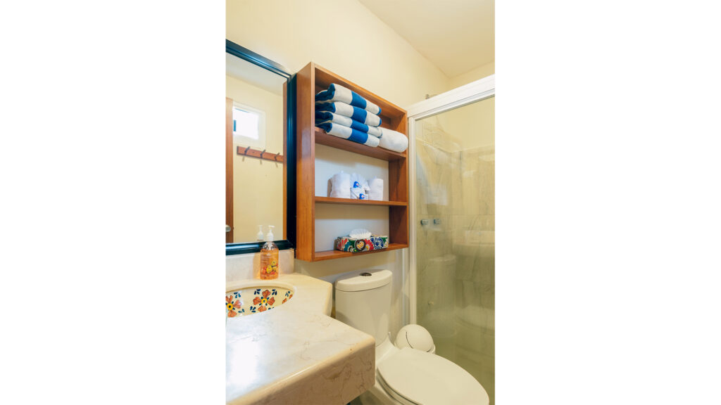 Bathroom vanity, toilet, and shower beside wooden shelves holding rolled towels.