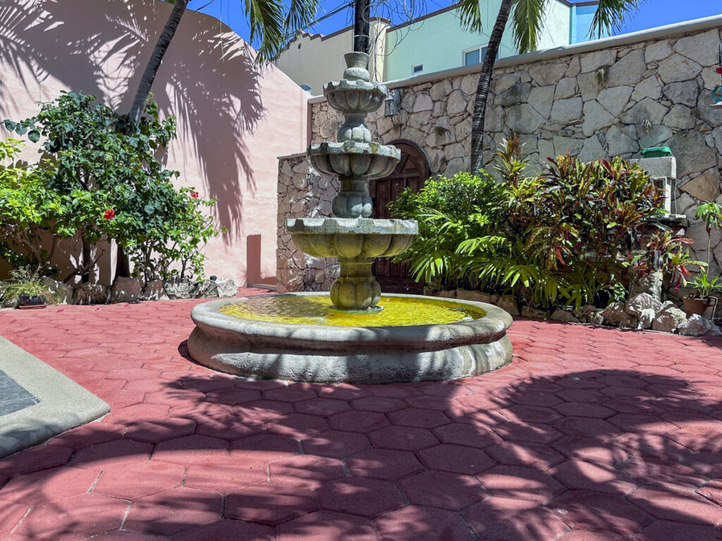 Large tiered stone fountain in a sunny courtyard with red paving and a stone boundary wall.