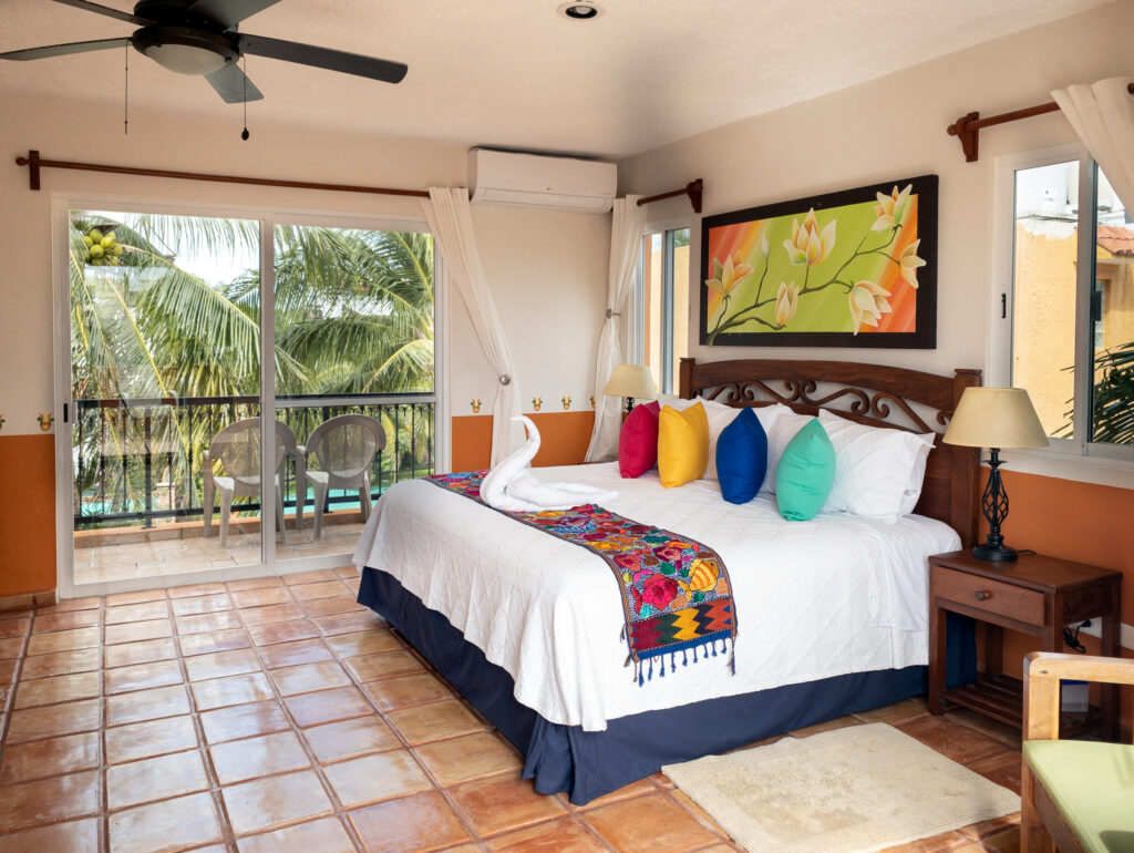 Bed with bright cushions beside glass doors and windows overlooking palm trees.
