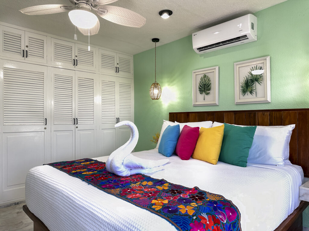 Bed with colorful pillows and floral runner beside white closet doors in a pale green bedroom.