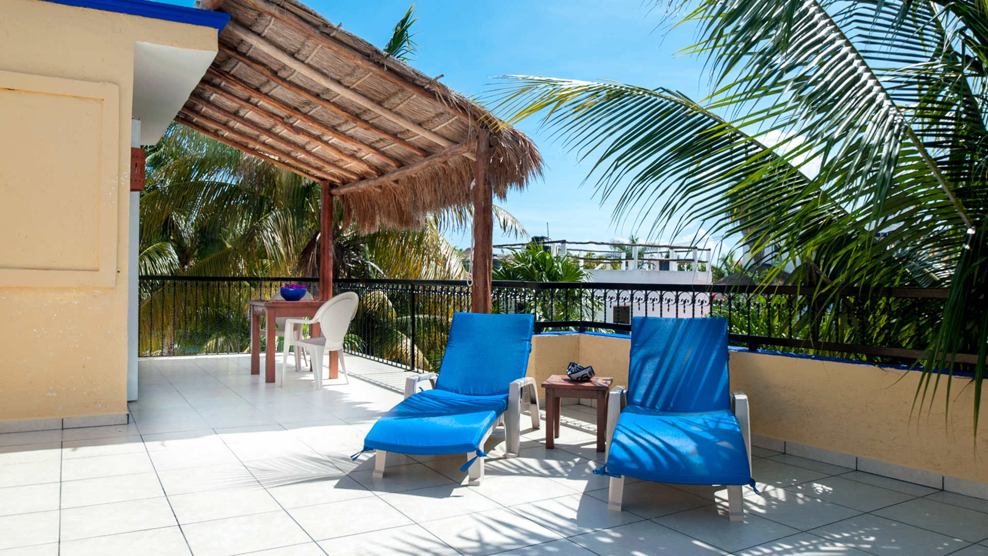 Two blue lounge chairs beside a small table on a broad terrace near a thatched shade structure.