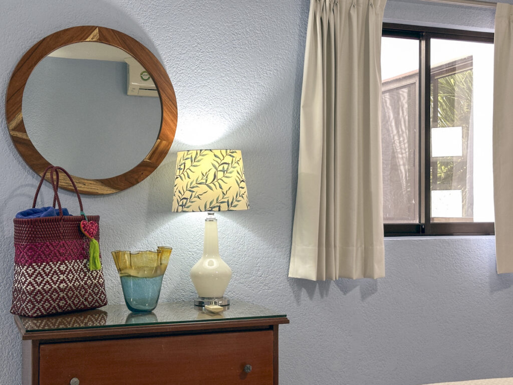 Wooden dresser with a patterned lamp, woven bag, and round mirror beside a curtained window.