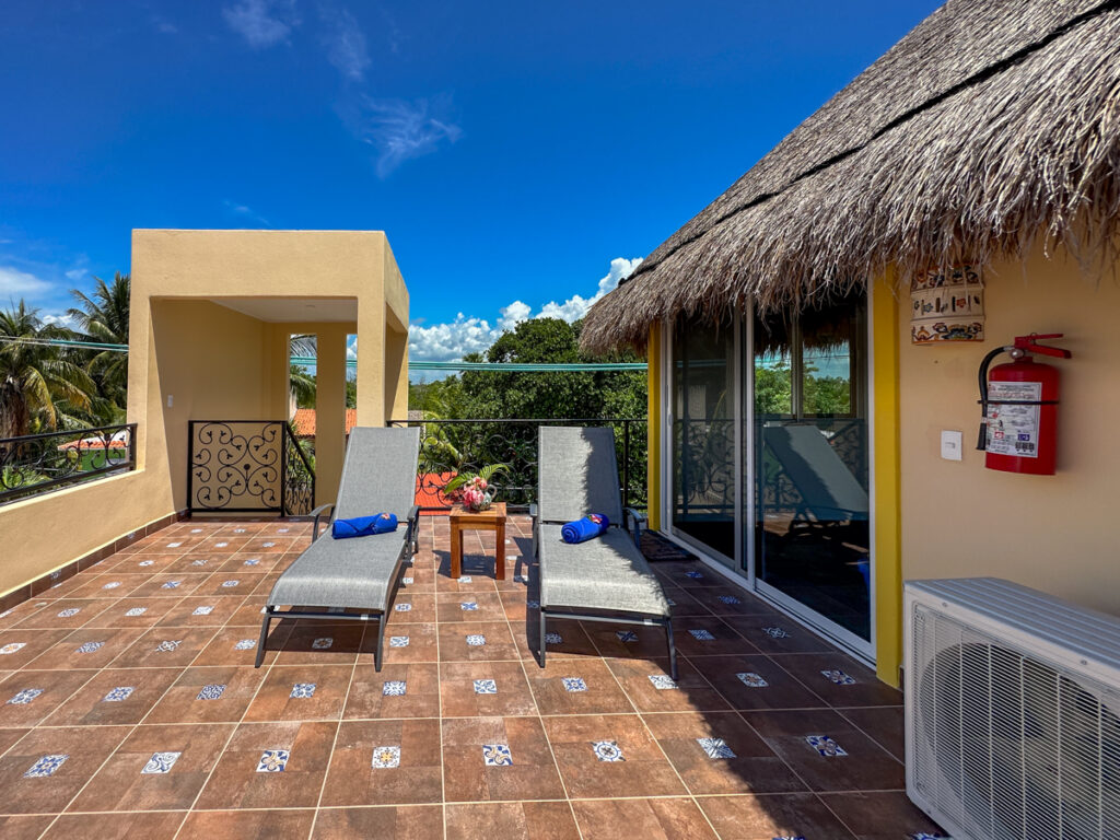 Two lounge chairs on a sunny tiled terrace beside a thatched roof and glass doors.