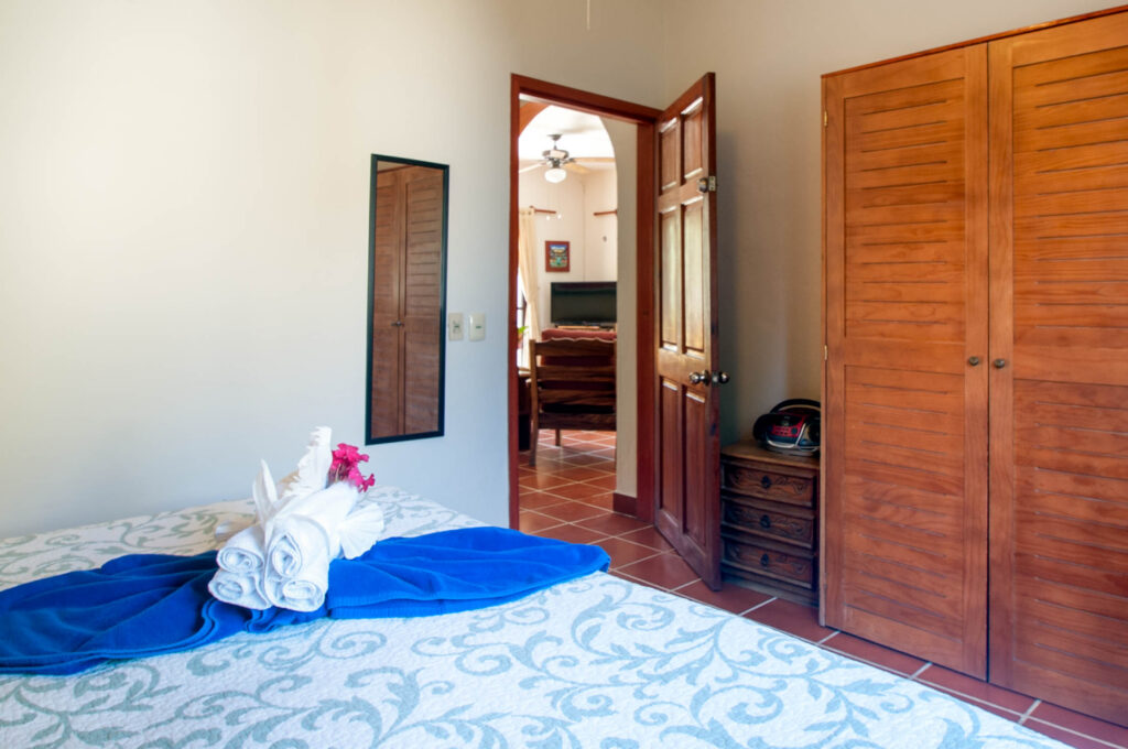 Bed with a blue towel and folded decoration faces an open doorway toward the dining area.