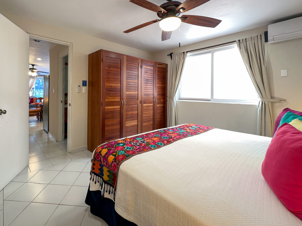 Bed with a floral runner beside a wooden wardrobe and curtained window.