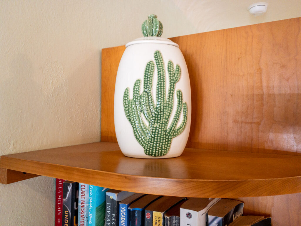 White ceramic jar decorated with a green cactus on a wooden shelf above books.