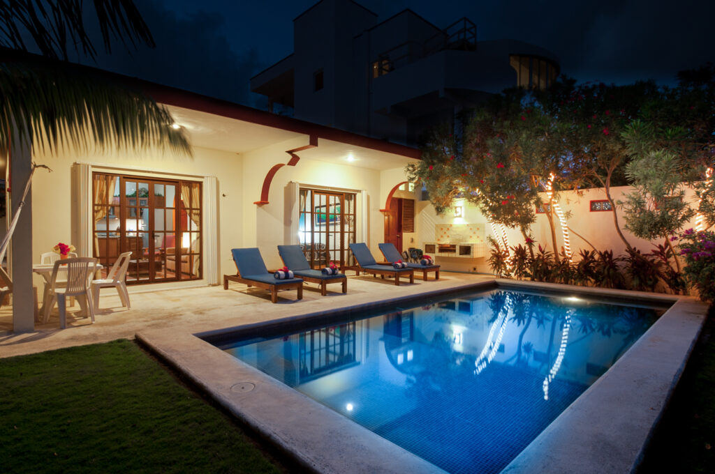 Lit pool in front of the house with lounge chairs and outdoor dining furniture after dark.