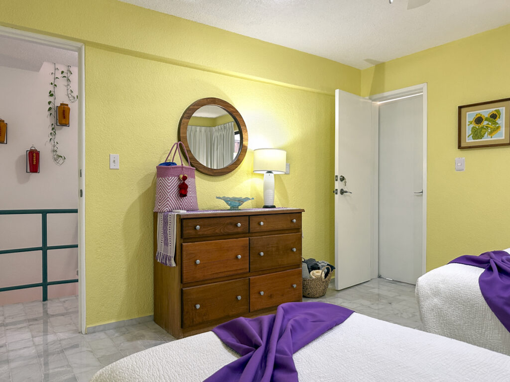 Wooden dresser beneath a round mirror beside an open doorway in a yellow bedroom.