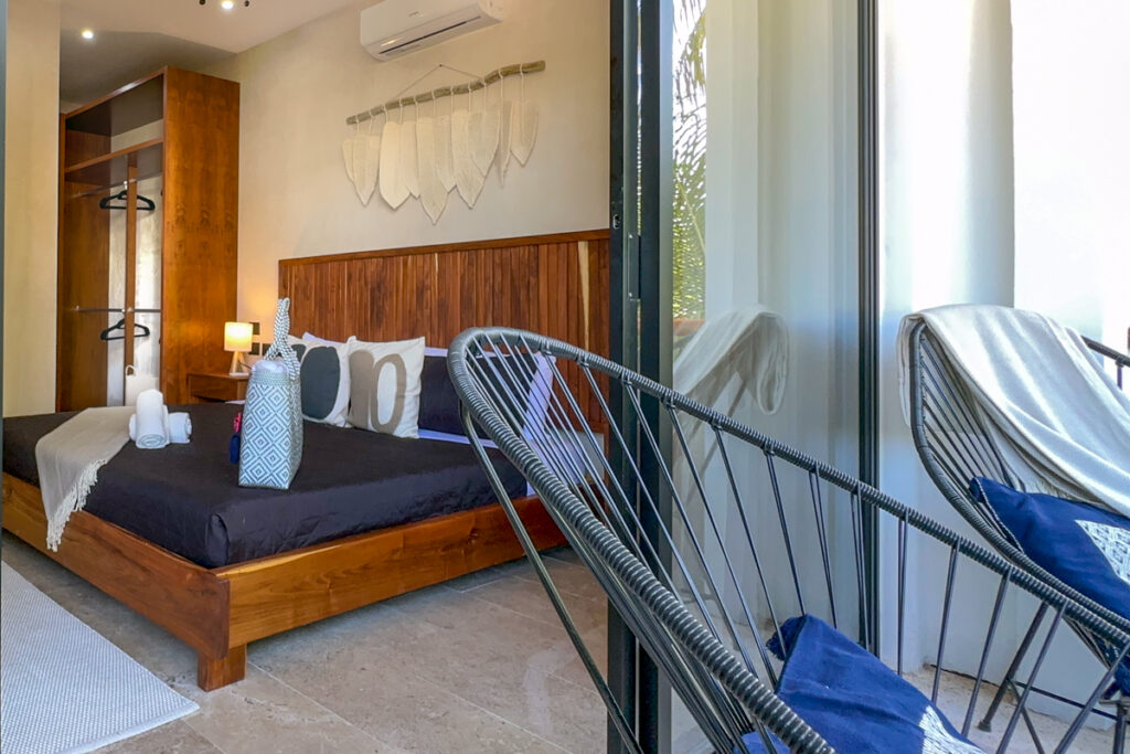 Wooden bed with dark bedding beside balcony chairs seen through an open sliding door.