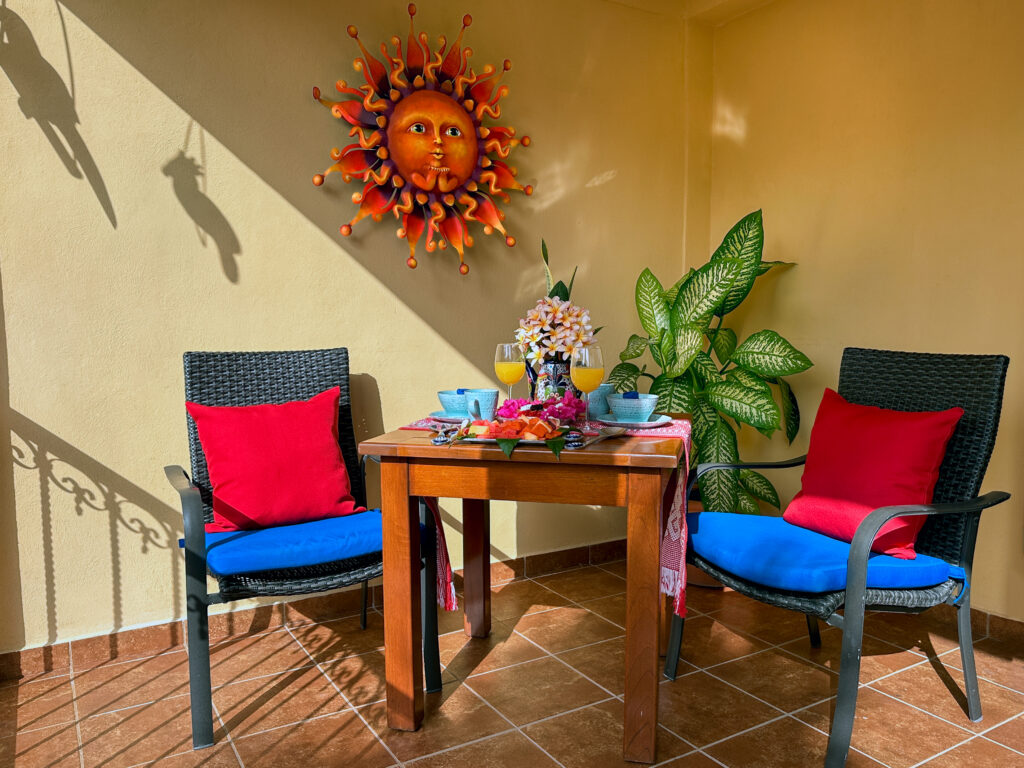 Two blue chairs with red cushions beside a small table beneath a colorful sun wall ornament.