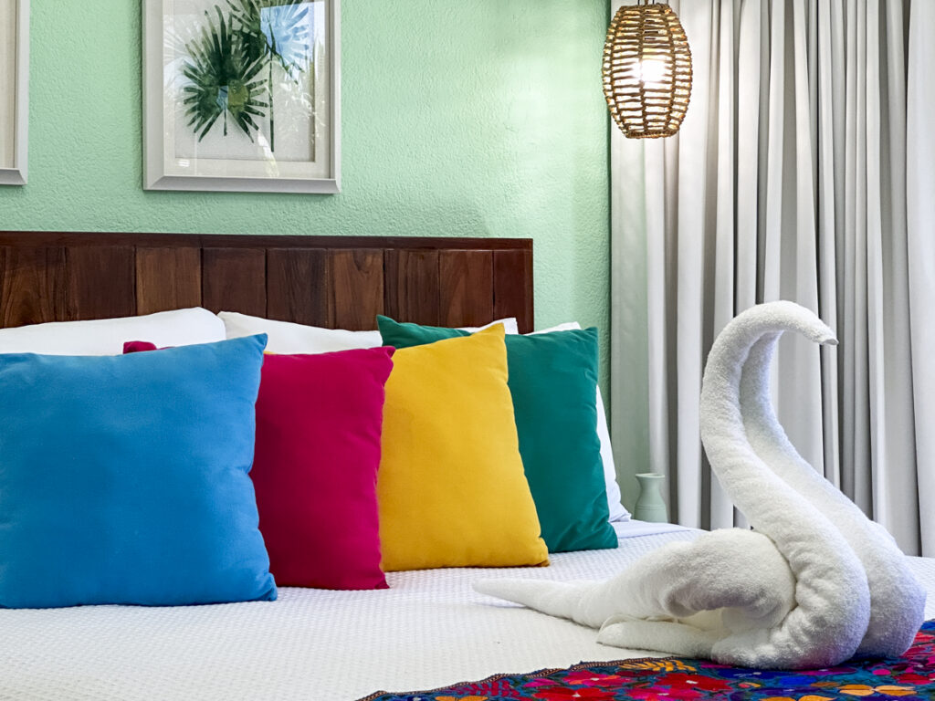 Blue, pink, yellow, and green pillows behind a white towel swan on a floral bed runner.
