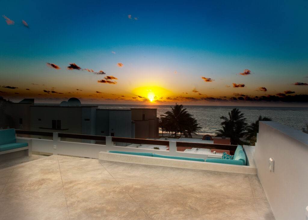 Low sun glows above the coastal horizon beyond rooftops and a white terrace railing.