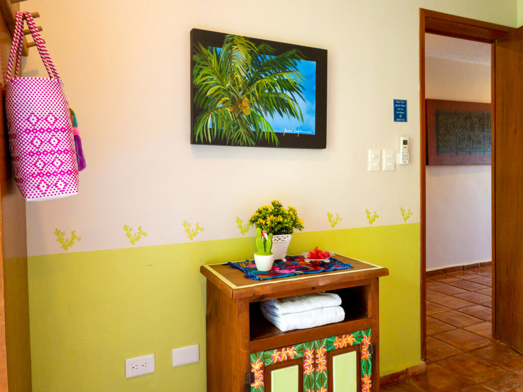 Small painted side table with flowers beneath palm artwork beside a hanging pink woven bag.
