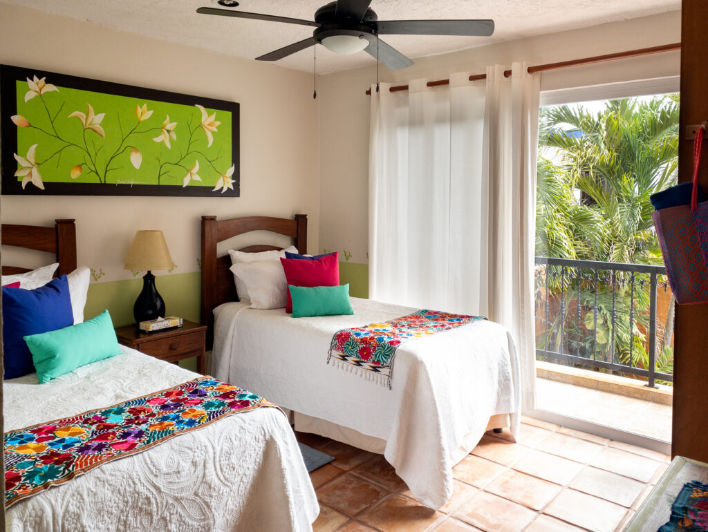 Two beds with bright bedding accents beside glass doors overlooking palms.