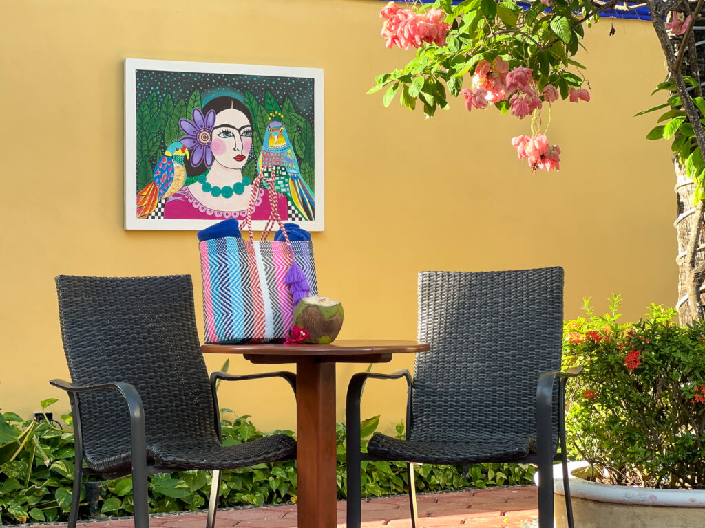 Two chairs and a wooden table beneath colorful portrait artwork on a yellow courtyard wall.