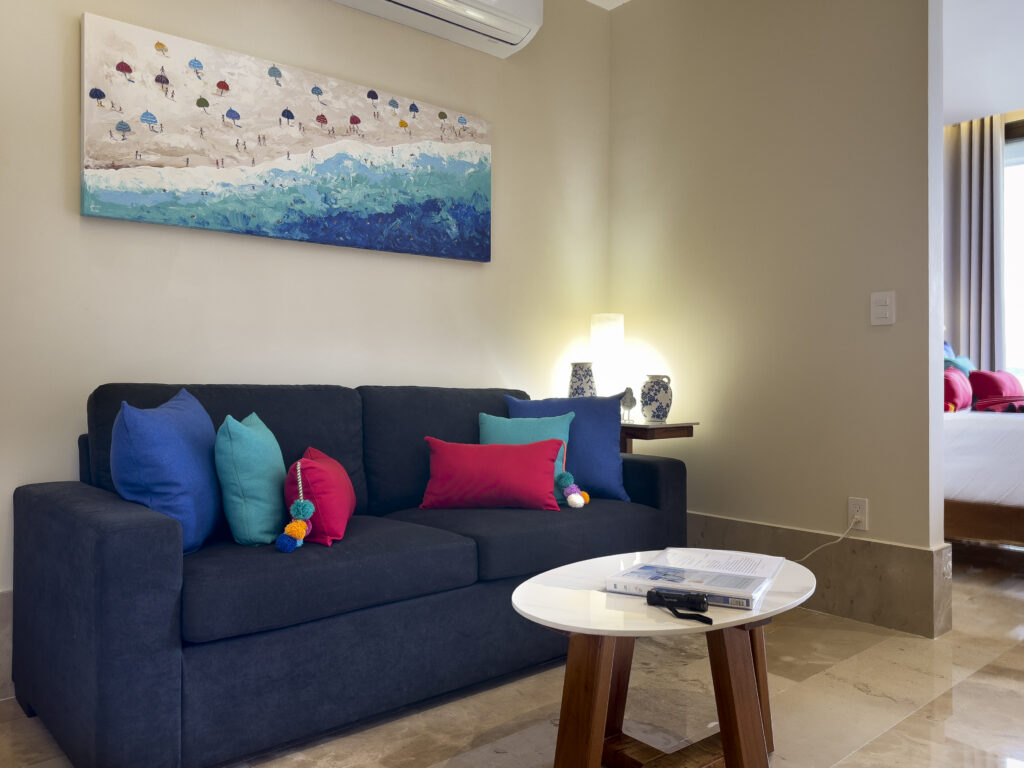 Dark sofa with colorful cushions beside a round coffee table beneath coastal artwork.