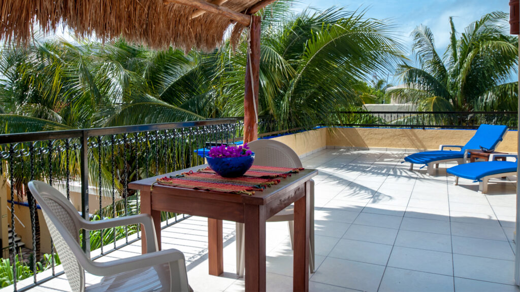 Small dining table beneath a thatched canopy beside terrace railings and palm trees.