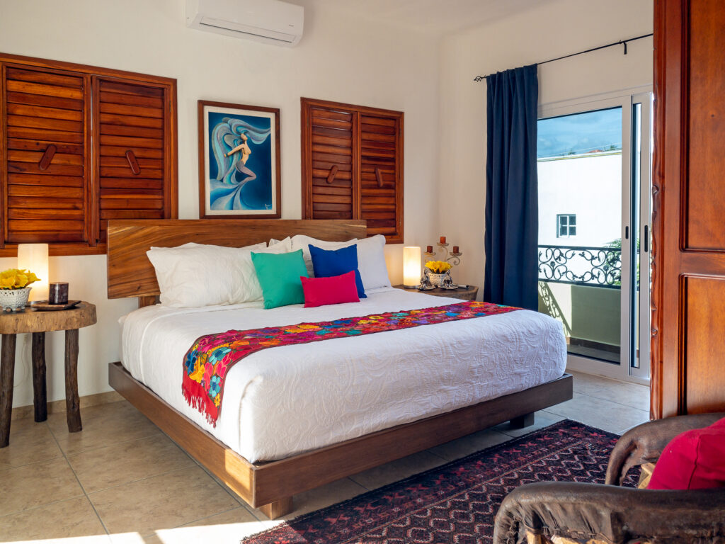 Bed with colorful accents and wooden headboard beneath framed artwork beside balcony doors.