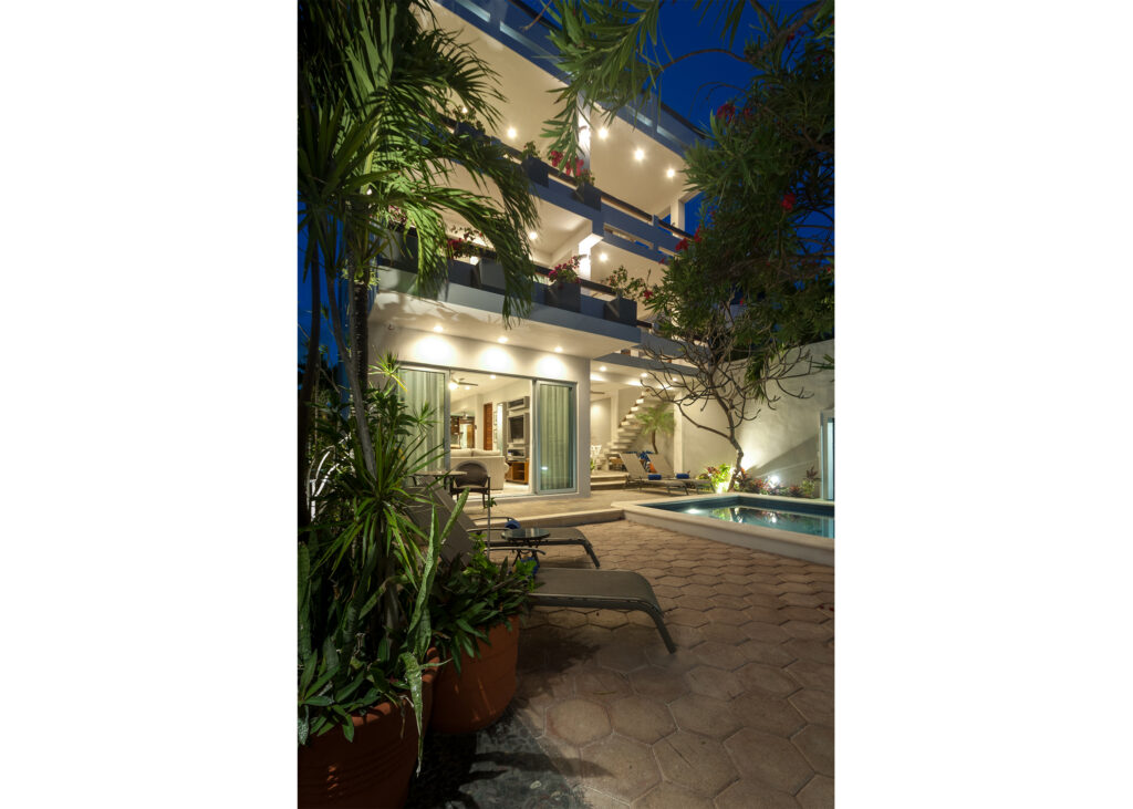 Lit house and pool courtyard seen along a garden path beneath palms at night.