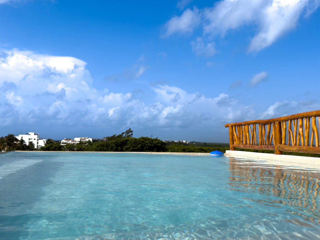 Wide rooftop pool beneath a blue sky with a wooden railing and treetops beyond.