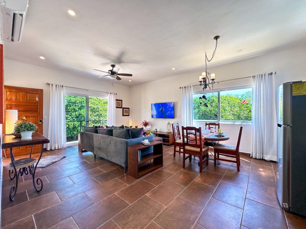 Wide living and dining room with a gray sectional, wooden table, television, and large windows.